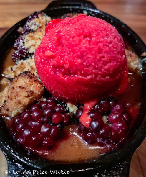 A photo of the berry cobbler which is made in small cast iron pots with ripe berries, snickerdoodle crumbs and topped with blackberry ice cream.